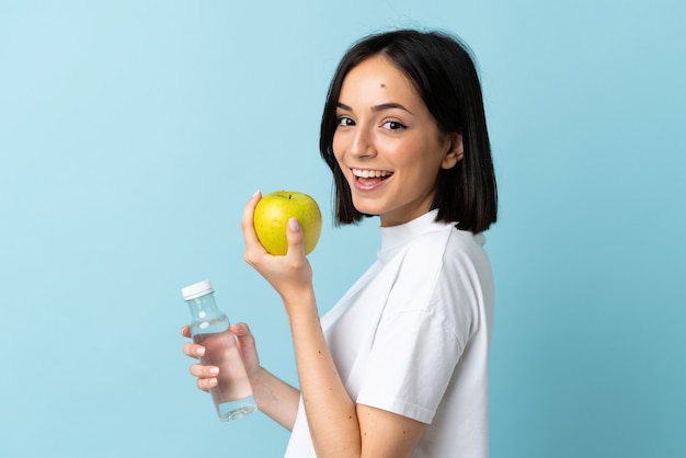 Young caucasian woman isolated on blue background with an apple and with a bottle of water