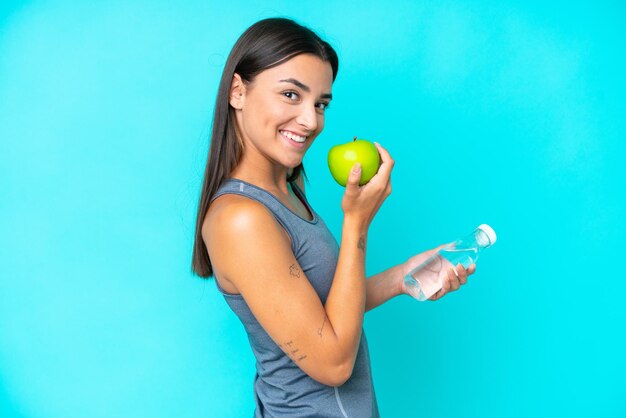 Young caucasian woman isolated on blue background with an apple and with a bottle of water