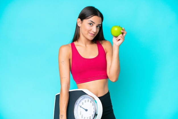 Young caucasian woman isolated on blue background with weighing machine and with an apple