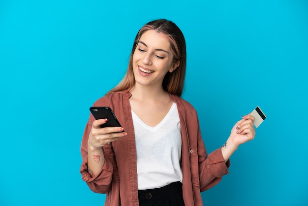 Young caucasian woman isolated on blue buying with the mobile with a credit card