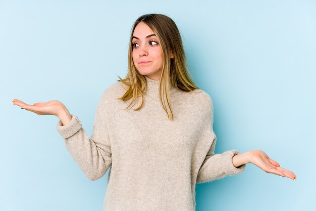 Young caucasian woman isolated on blue confused and doubtful shrugging shoulders to hold a copy space.