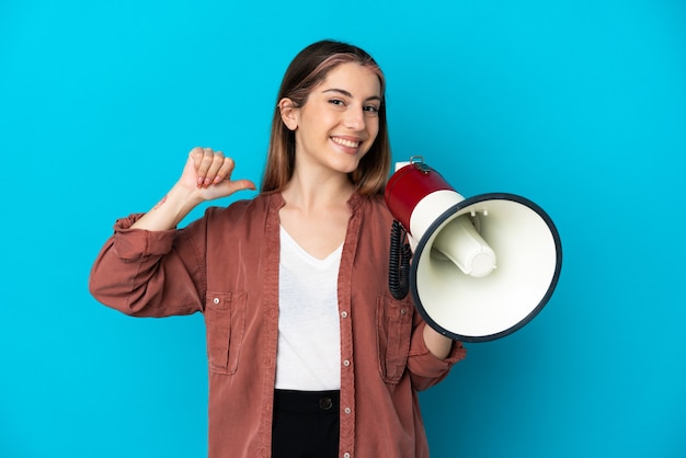 Young caucasian woman isolated on blue holding a megaphone and proud and self-satisfied