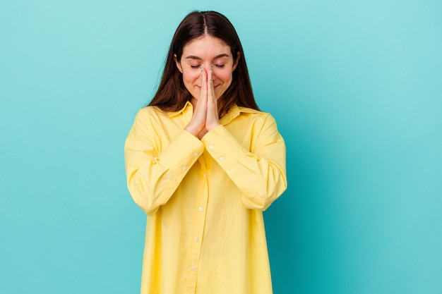 Young caucasian woman isolated on blue wall holding hands in pray near mouth, feels confident.