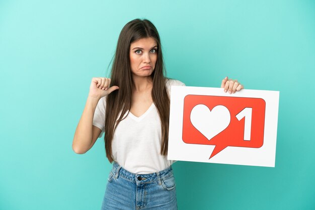 Young caucasian woman isolated on blue wall holding a placard with Like icon with proud gesture