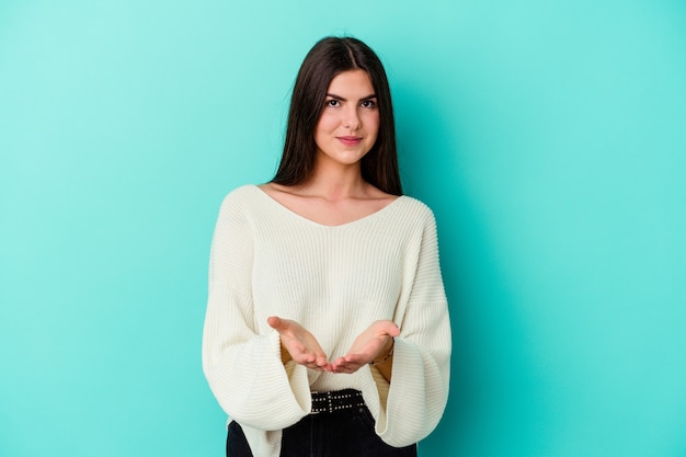 Young caucasian woman isolated on blue wall holding something with palms, offering