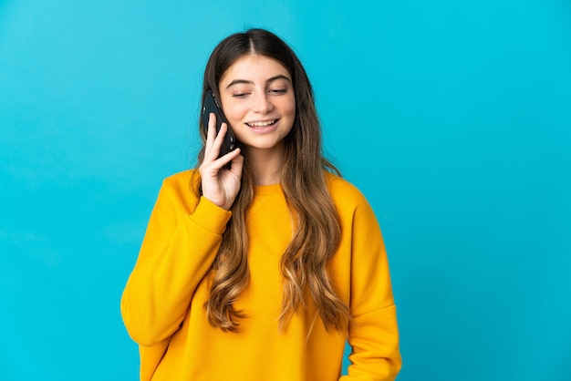 Young caucasian woman isolated on blue wall keeping a conversation with the mobile phone with someone