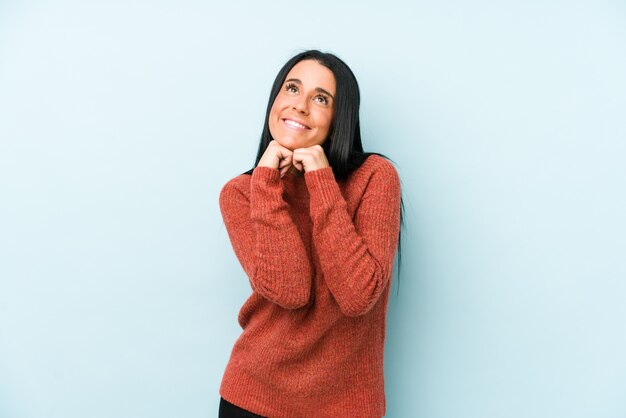 Young caucasian woman isolated on a blue wall keeps hands under chin, is looking happily aside.