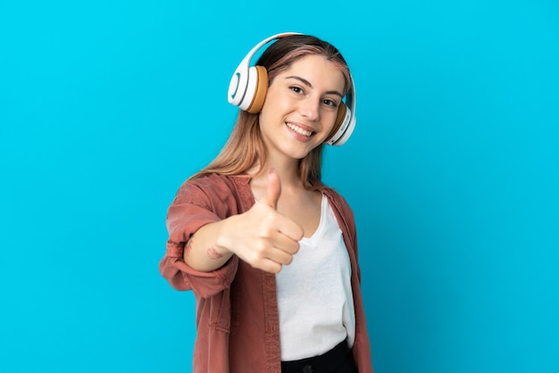 Young caucasian woman isolated on blue wall listening music and with thumb up