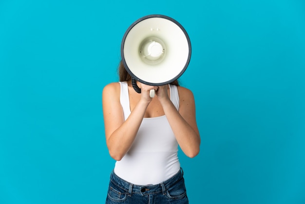 Young caucasian woman isolated on blue wall shouting through a megaphone to announce something