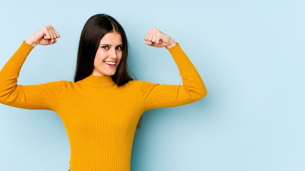 Photo young caucasian woman isolated on blue wall showing strength gesture with arms, symbol of feminine power