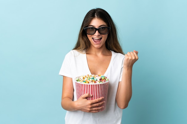 Young caucasian woman isolated on blue with 3d glasses and holding a big bucket of popcorns