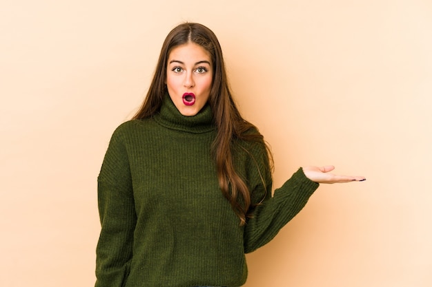 Young caucasian woman isolated en beige impressed holding copy space on palm.