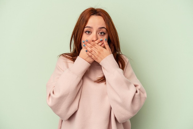 Young caucasian woman isolated on green background covering mouth with hands looking worried.