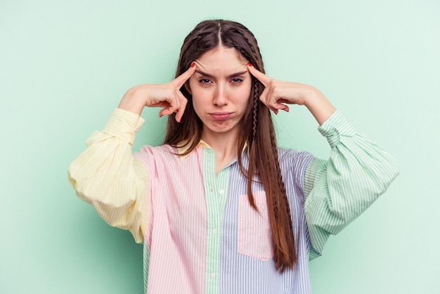 Young caucasian woman isolated on green background focused on a task keeping forefingers pointing head