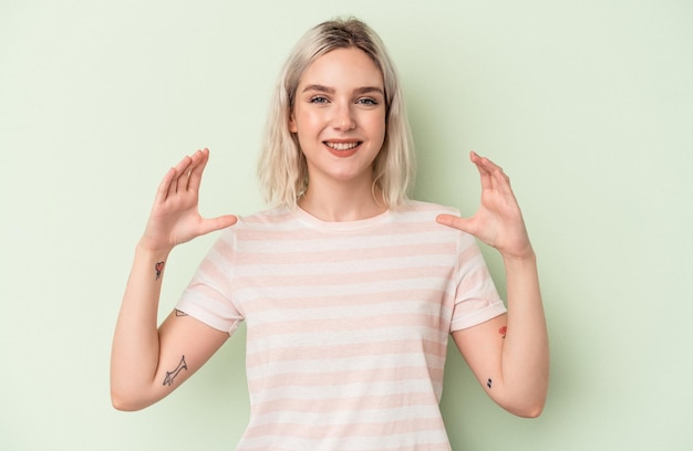 Young caucasian woman isolated on green background holding something with palms, offering to camera.