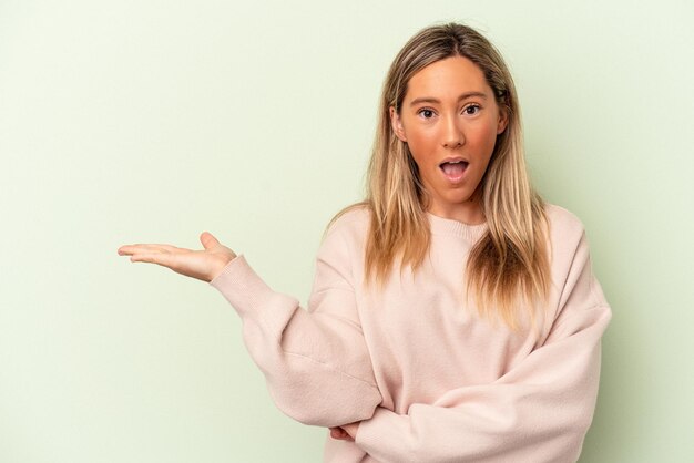 Young caucasian woman isolated on green background impressed holding copy space on palm.