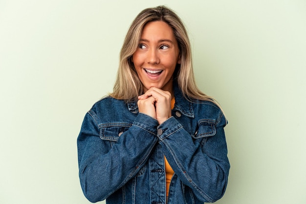 Young caucasian woman isolated on green background keeps hands under chin, is looking happily aside.