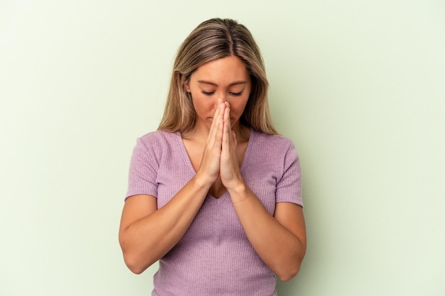 Young caucasian woman isolated on green background praying, showing devotion, religious person looking for divine inspiration.