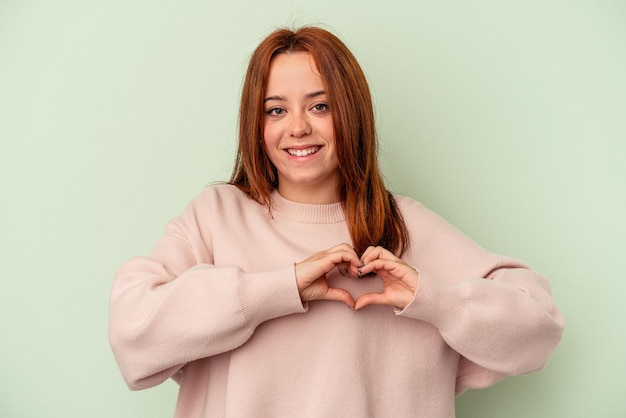 Young caucasian woman isolated on green background smiling and showing a heart shape with hands.