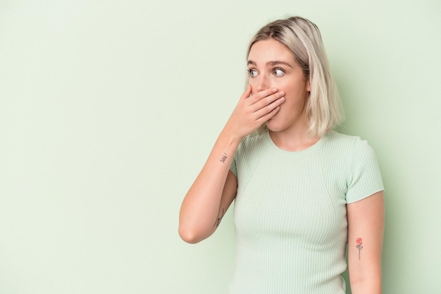 Young caucasian woman isolated on green background thoughtful looking to a copy space covering mouth with hand.