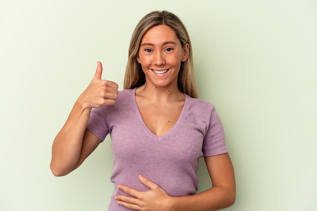 Young caucasian woman isolated on green background touches tummy, smiles gently, eating and satisfaction concept.