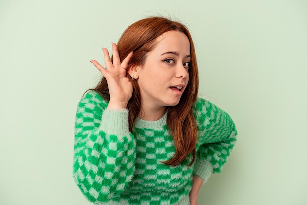 young caucasian woman isolated on green background trying to listening a gossip.
