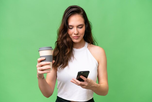 Young caucasian woman isolated over isolated background holding coffee to take away and a mobile