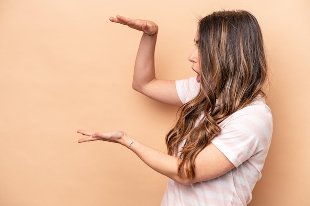 写真 ベージュの背景に孤立した若い白人女性は、手の間にコピースペースを持ってショックを受けて驚いています