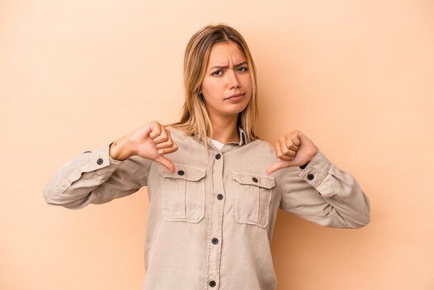 写真 親指を下に、失望の概念を示すベージュの背景に分離された若い白人女性。