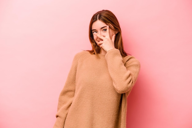 Young caucasian woman isolated on pink background blink at the camera through fingers embarrassed covering face