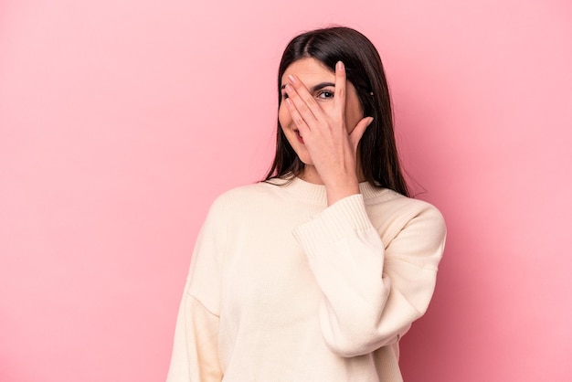 Young caucasian woman isolated on pink background blink at the camera through fingers embarrassed covering face