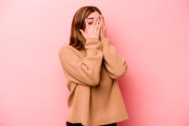 Young caucasian woman isolated on pink background blink through fingers frightened and nervous
