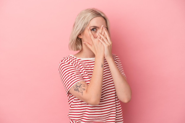 Young caucasian woman isolated on pink background blink through fingers frightened and nervous