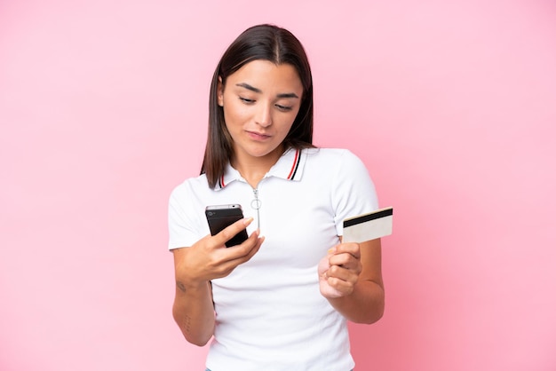 Молодая кавказская женщина изолирована на розовом фоне, покупая с мобильного телефона с помощью кредитной карты