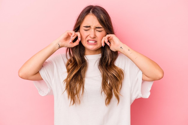 Young caucasian woman isolated on pink background covering ears with fingers, stressed and desperate by a loudly ambient.