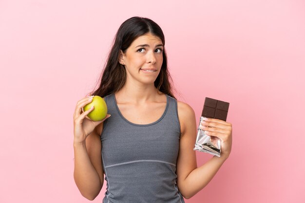 Young caucasian woman isolated on pink background having doubts while taking a chocolate tablet in one hand and an apple in the other