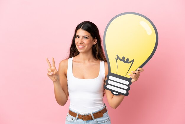 Foto giovane donna caucasica isolata su sfondo rosa che tiene in mano un'icona a forma di lampadina e celebra una vittoria