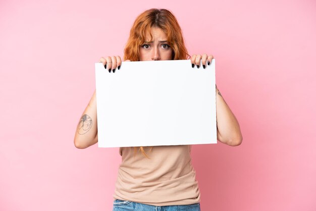 분홍색 배경 에 고립 된 젊은 백인 여성 은 빈 표지판 을 들고 그 뒤 에 숨어 있다