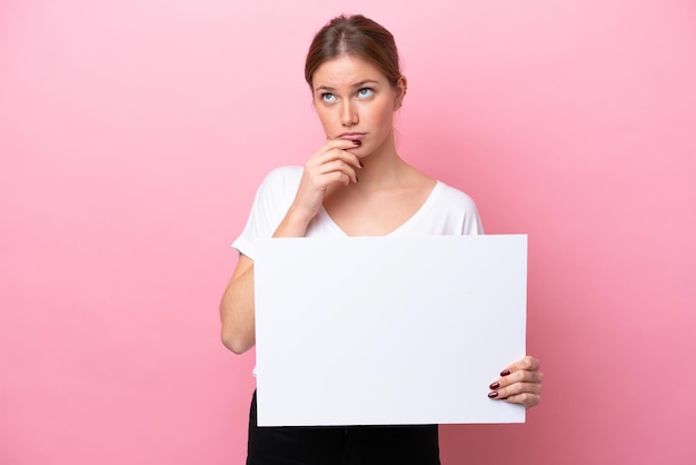 Young caucasian woman isolated on pink background holding an empty placard and thinking