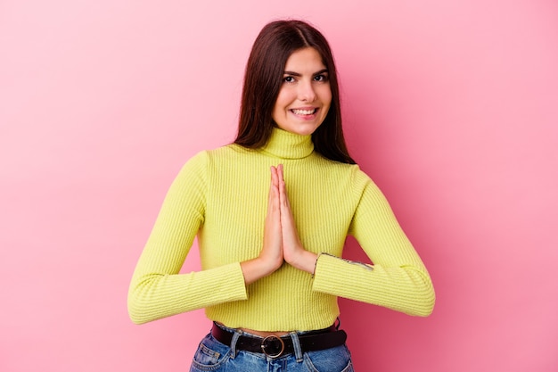 Young caucasian woman isolated on pink background holding hands in pray near mouth, feels confident.