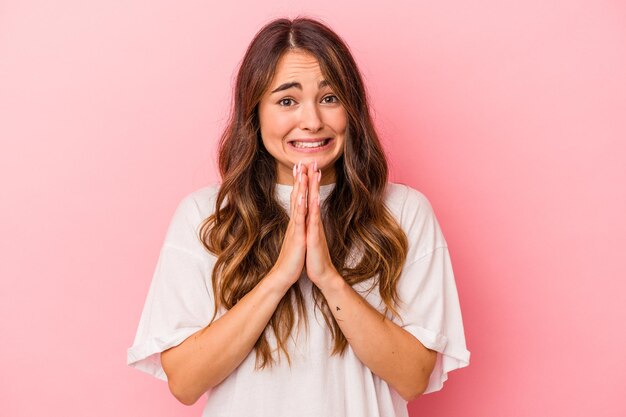 Young caucasian woman isolated on pink background holding hands in pray near mouth, feels confident.