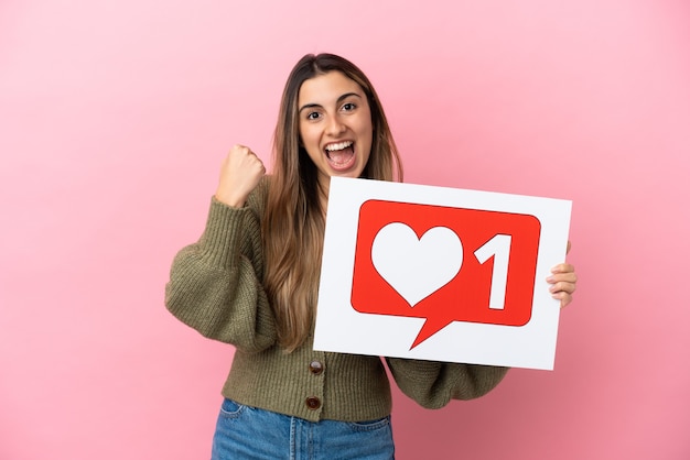 Young caucasian woman isolated on pink background holding a placard with Like icon and celebrating a victory