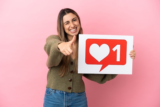 Photo young caucasian woman isolated on pink background holding a placard with like icon and pointing to the front