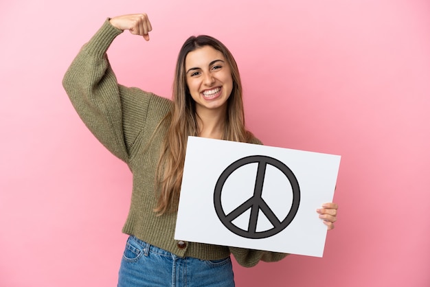 Young caucasian woman isolated on pink background holding a placard with peace symbol and doing strong gesture