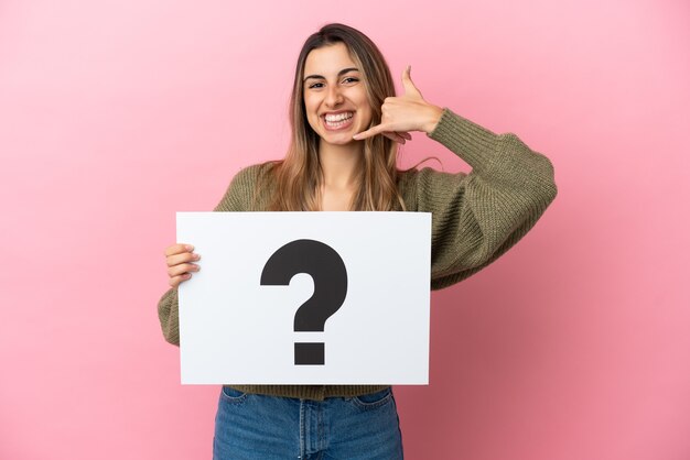 Young caucasian woman isolated on pink background holding a placard with question mark symbol and doing phone gesture