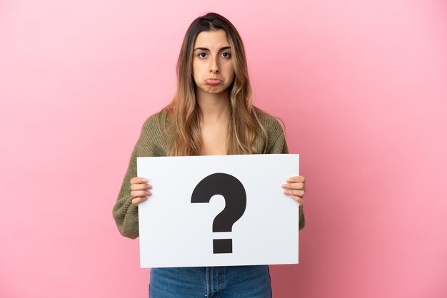 Young caucasian woman isolated on pink background holding a placard with question mark symbol with sad expression