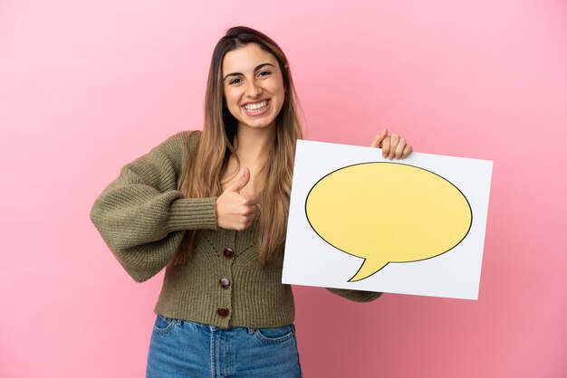 Young caucasian woman isolated on pink background holding a placard with speech bubble icon with thumb up