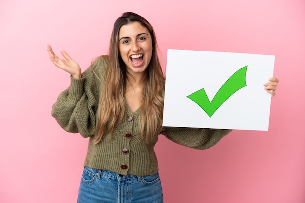 Young caucasian woman isolated on pink background holding a placard with text Green check mark icon with surprised expression