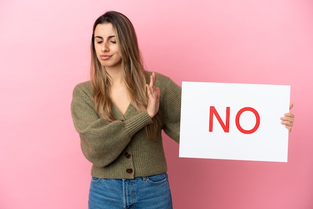 Giovane donna caucasica isolata su sfondo rosa, tenendo un cartello con il testo no e facendo il segnale di stop