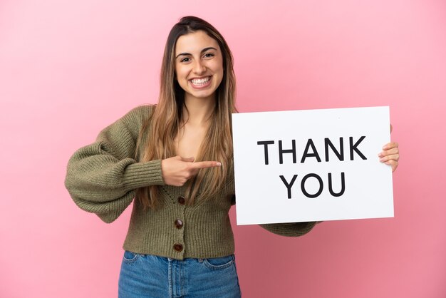 Young caucasian woman isolated on pink background holding a placard with text THANK YOU and  pointing it